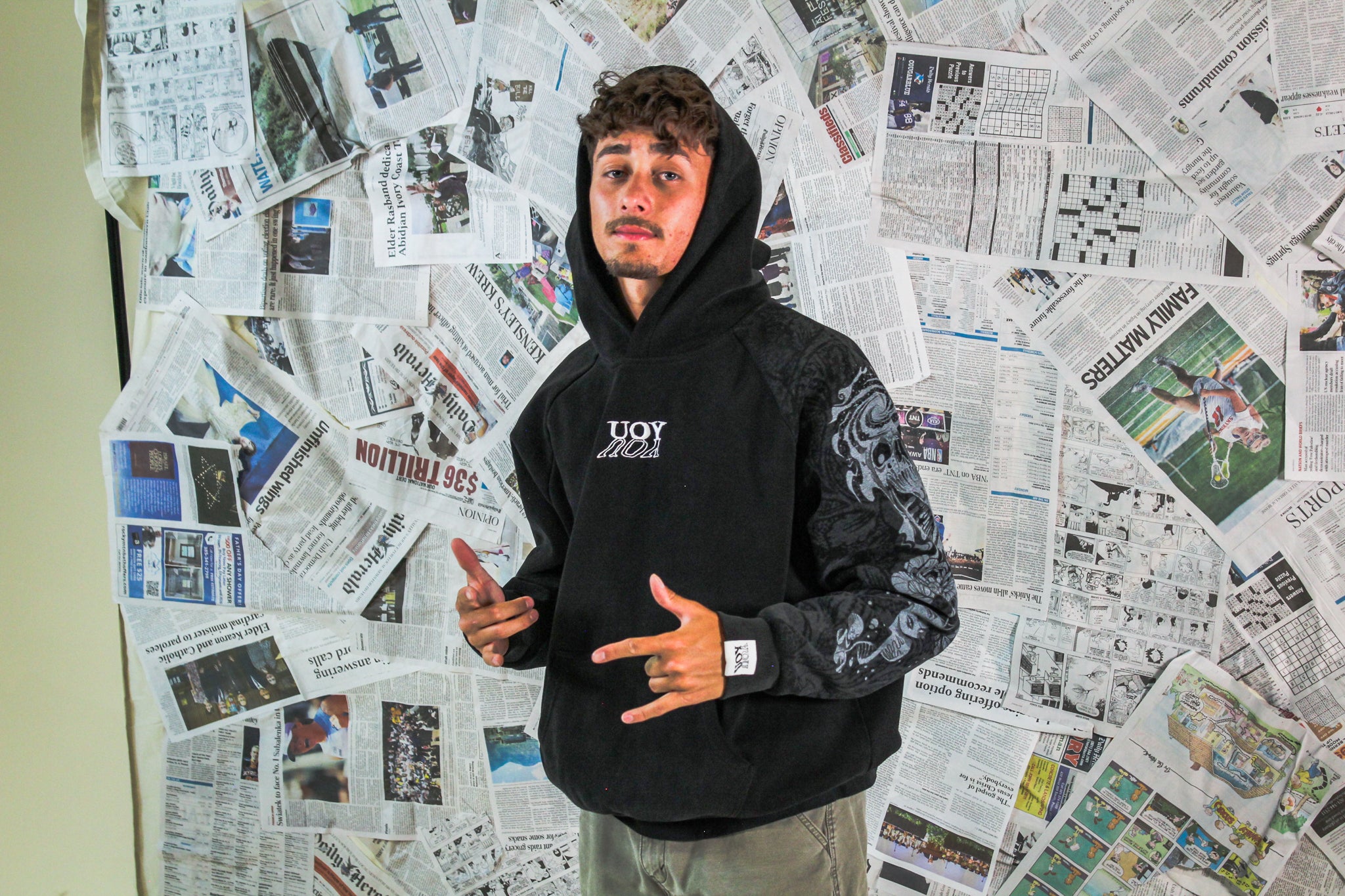 Person wearing a black hoodie with a logo, standing in front of a wall covered with newspapers.