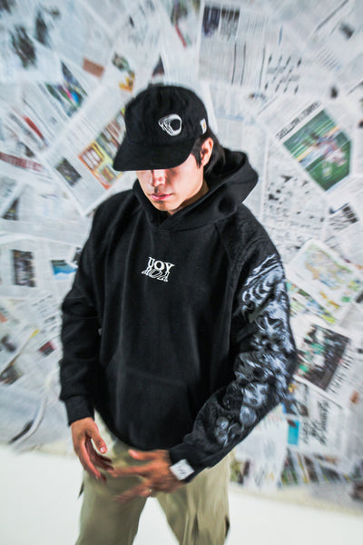 Person wearing a black hoodie with a logo and a black cap in front of a newspaper wall.