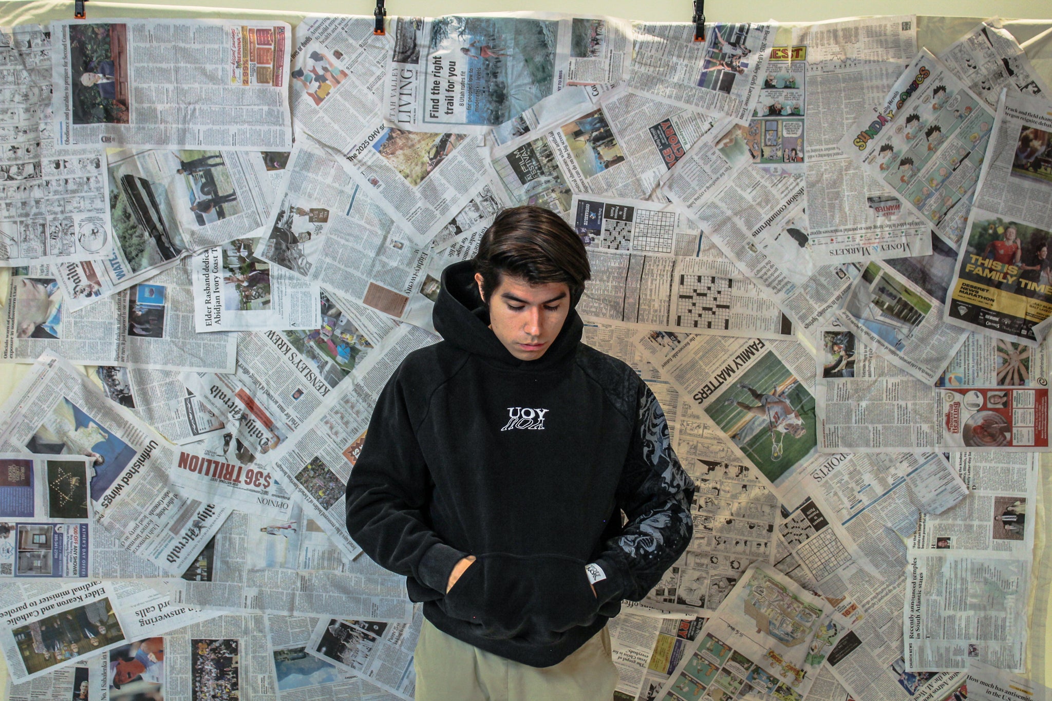Person wearing a black hoodie with visible branding standing amidst a collage of newspapers.