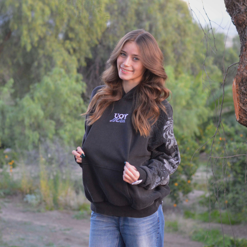 Woman wearing a black hoodie with a logo, standing outdoors near trees.