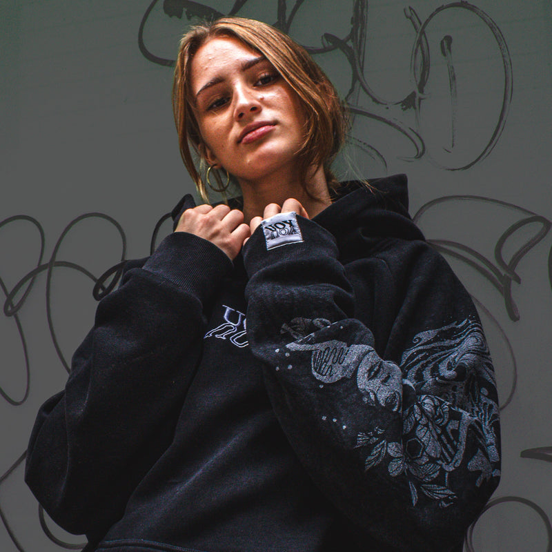 Person wearing a YOU hoodie with a brand logo, standing against a graffiti-covered wall.