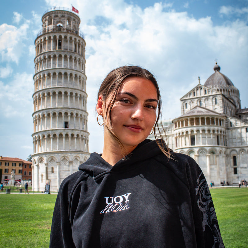 Woman standing in front of the Leaning Tower of Pisa with a clear blue sky. Wearing a YOU hoodie