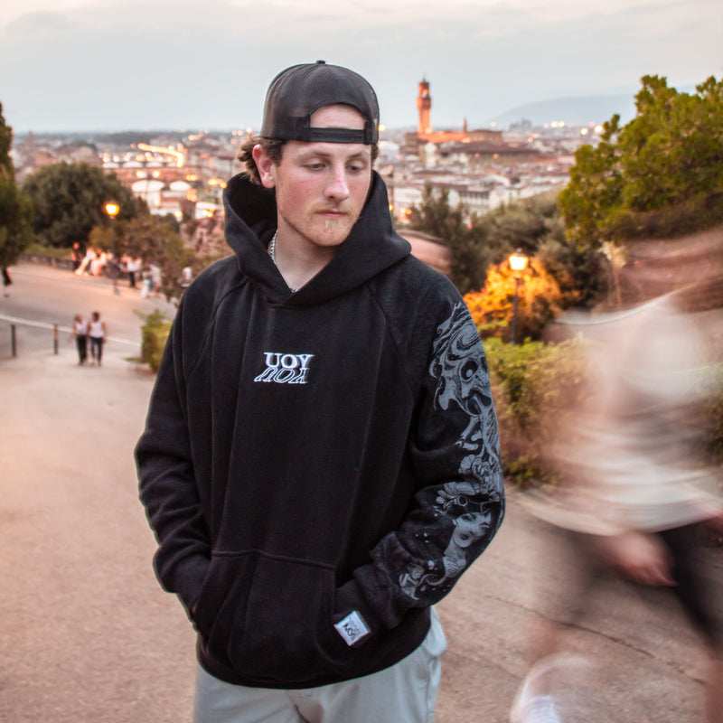 Person wearing a YOU hoodie with a logo, standing outdoors with a cityscape in the background.