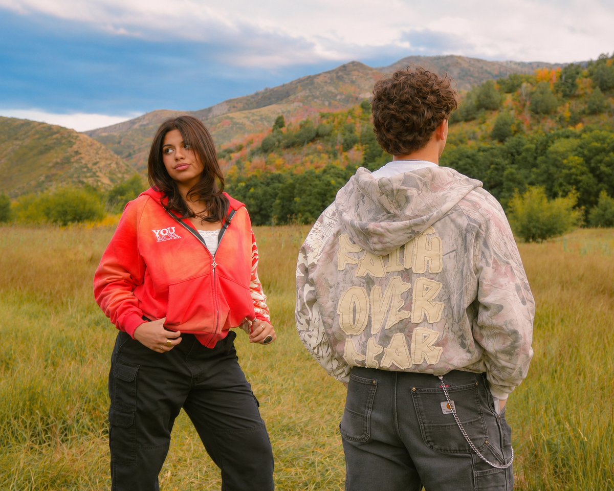 Two models standing in an outdoor grassy field with mountains and autumn trees in the background. The woman on the left wears a bright coral-red zip-up hoodie with 'FOY' printed on the left chest and black cargo pants.