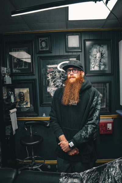 Man with a beard and glasses standing in a tattoo parlor with framed artwork on the wall.