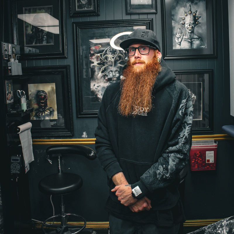Man with a beard and glasses standing in a tattoo parlor with framed artwork on the wall.