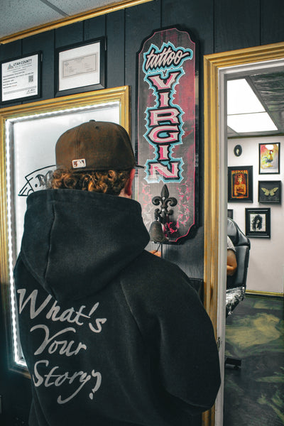 Person wearing a hoodie with text, standing in front of a tattoo parlor sign.
